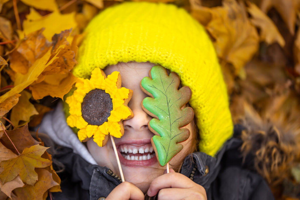 una ragazzina divertente con un cappello giallo giace nel fogliame autunnale e tiene mano il pan di zenzero